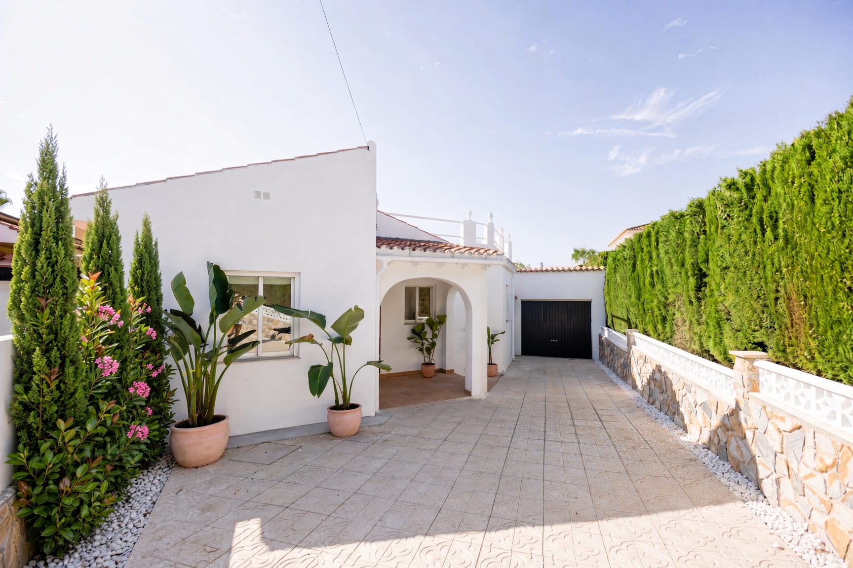 Courtyard driveway with a tall dense privacy hedge above the low stone wall on the left, white villa and dark garage door beyond